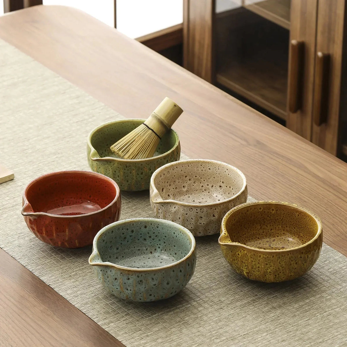 Five spouted matcha bowls in yellow, green, red, blue and white glazes on a table, with a bamboo whisk in the green bowl