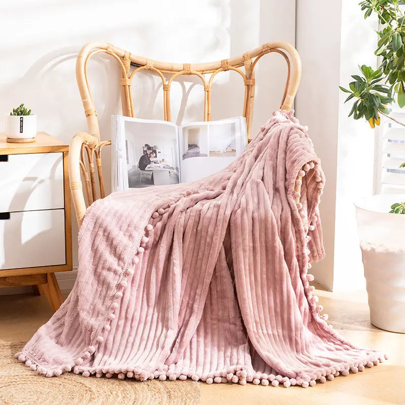 A pink flannel throw with raised stripes and a matching pom-pom edge falling from a rattan armchair to the floor