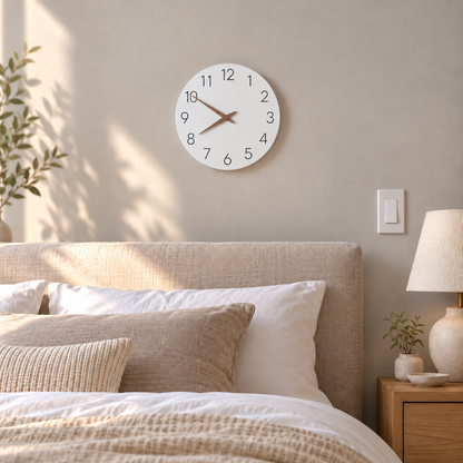 A small white wall clock with wooden hands above a linen headboard in a calm bedroom in the morning