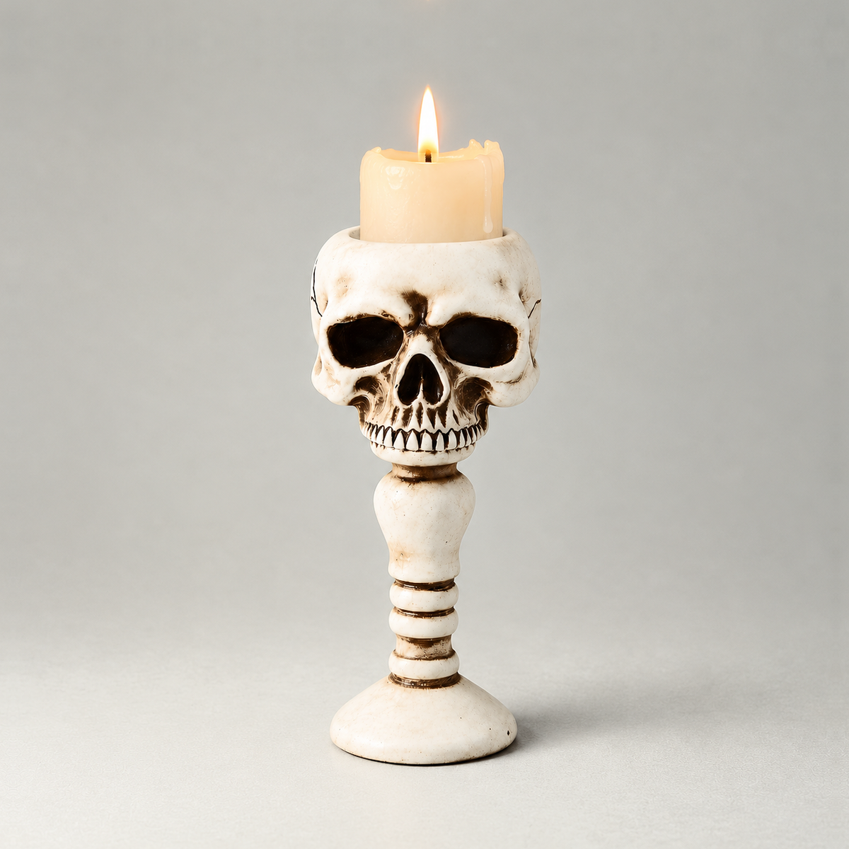 Bone skull candle holder shown front on against a plain grey studio background, with a short lit pillar candle standing in the well in the top of the head