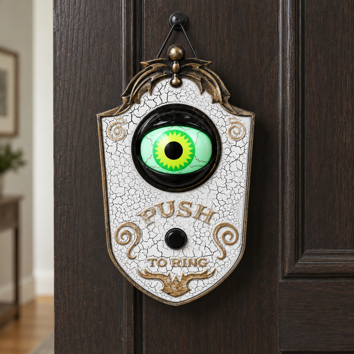White Halloween eyeball doorbell decoration with a crackle finish and a black socket ring, hanging on a door with a daylit hallway behind it, its single green eye lit