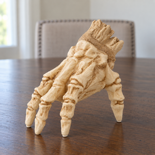 Unlit skeleton hand standing on its fingertips on a dark walnut dining table in daylight, with a pale upholstered dining chair behind it and a torn stump at the wrist