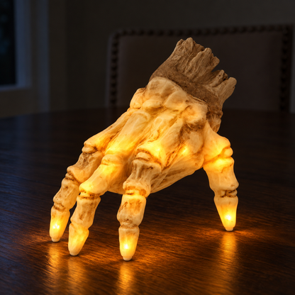 The skeleton hand lit on a dark walnut dining table at night, glowing warm amber from inside the bone at the knuckles and fingertips, its light pooling on the table, with the wrist stump dark