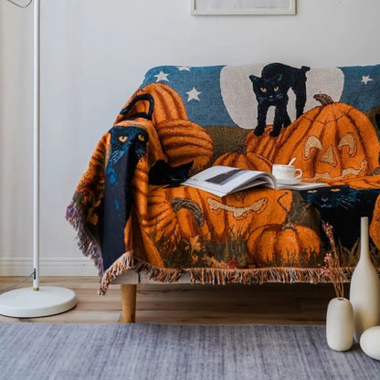 Woven Halloween throw blanket over a two-seat sofa in daylight, showing carved pumpkins, black cats and a full moon on a deep blue night sky, with a knotted tassel fringe along the lower edge
