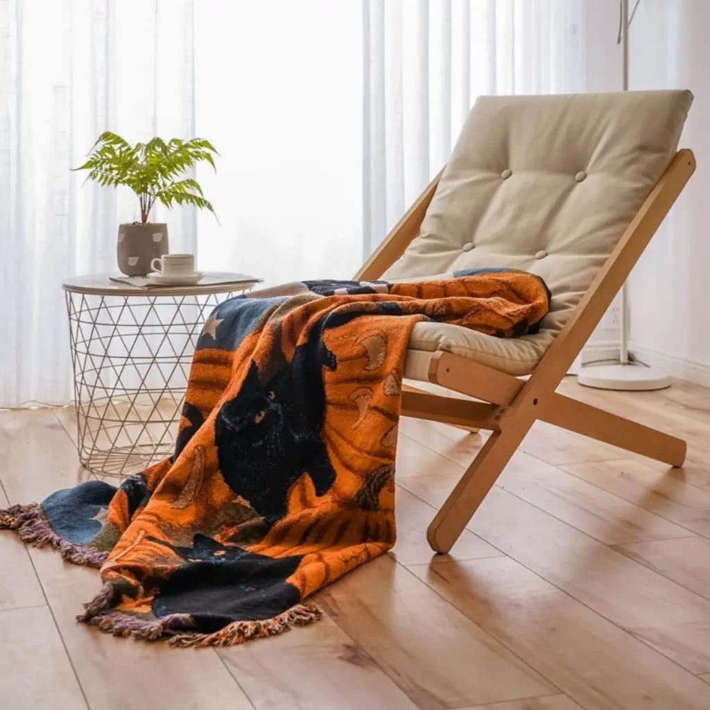 Pumpkin and black cat throw blanket draped over a wooden folding chair and trailing onto a wood floor, with the knotted tassel fringe visible along the edge