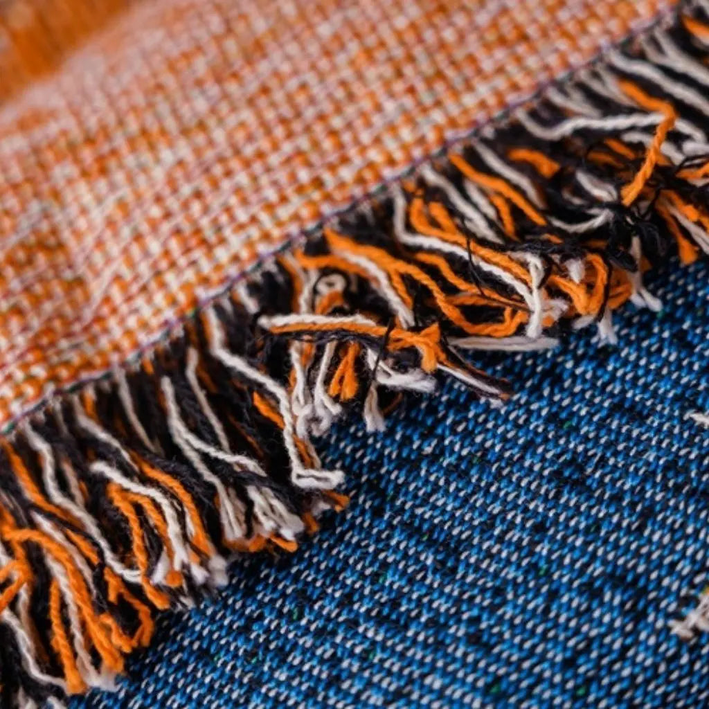 Macro of the blanket's edge, the stitched hem above and the knotted tassel fringe in orange, black and cream below it