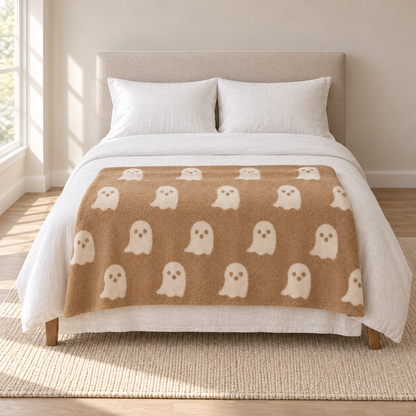 Ghost knit throw blanket in tan with ivory ghosts, spread across the foot of a made bed in morning daylight