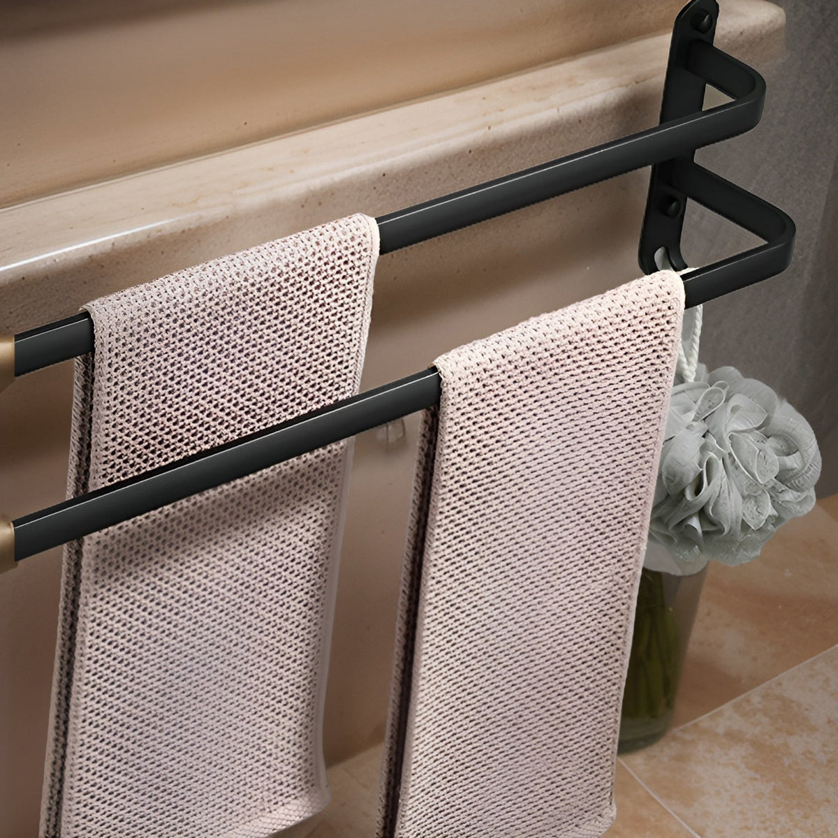 Two-bar matte black aluminum towel rack mounted below the rim of a bath, a towel on each stepped bar and a loofah hanging from a hook at the foot of the end plate
