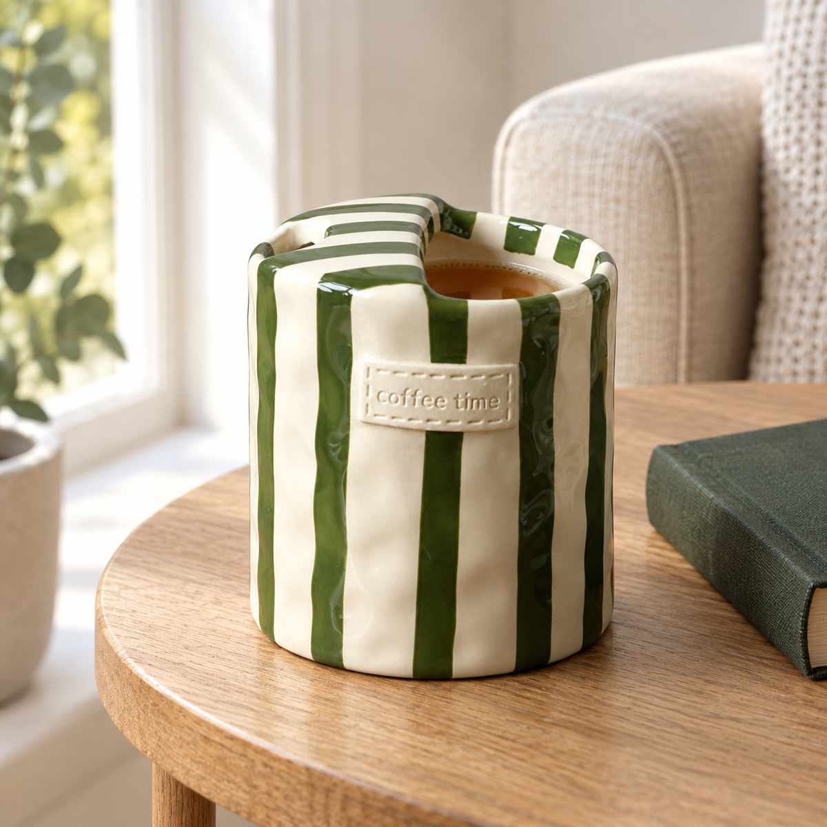 Green striped ceramic cup of tea on an oak side table beside a book and a linen armchair
