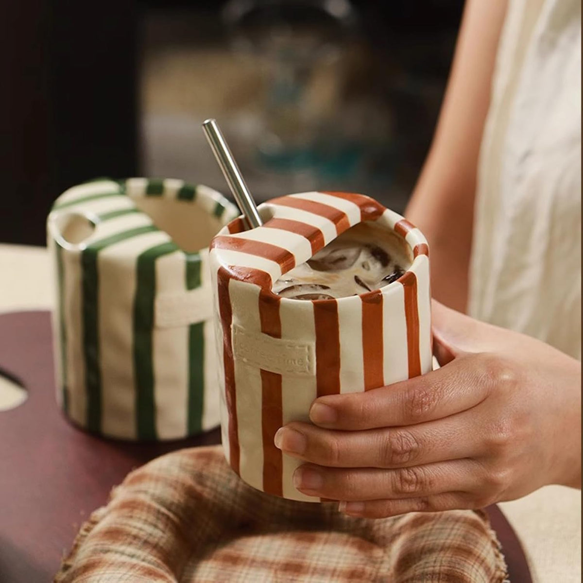 Brown striped ceramic cup of iced coffee with a steel straw through its folded top, a green cup behind