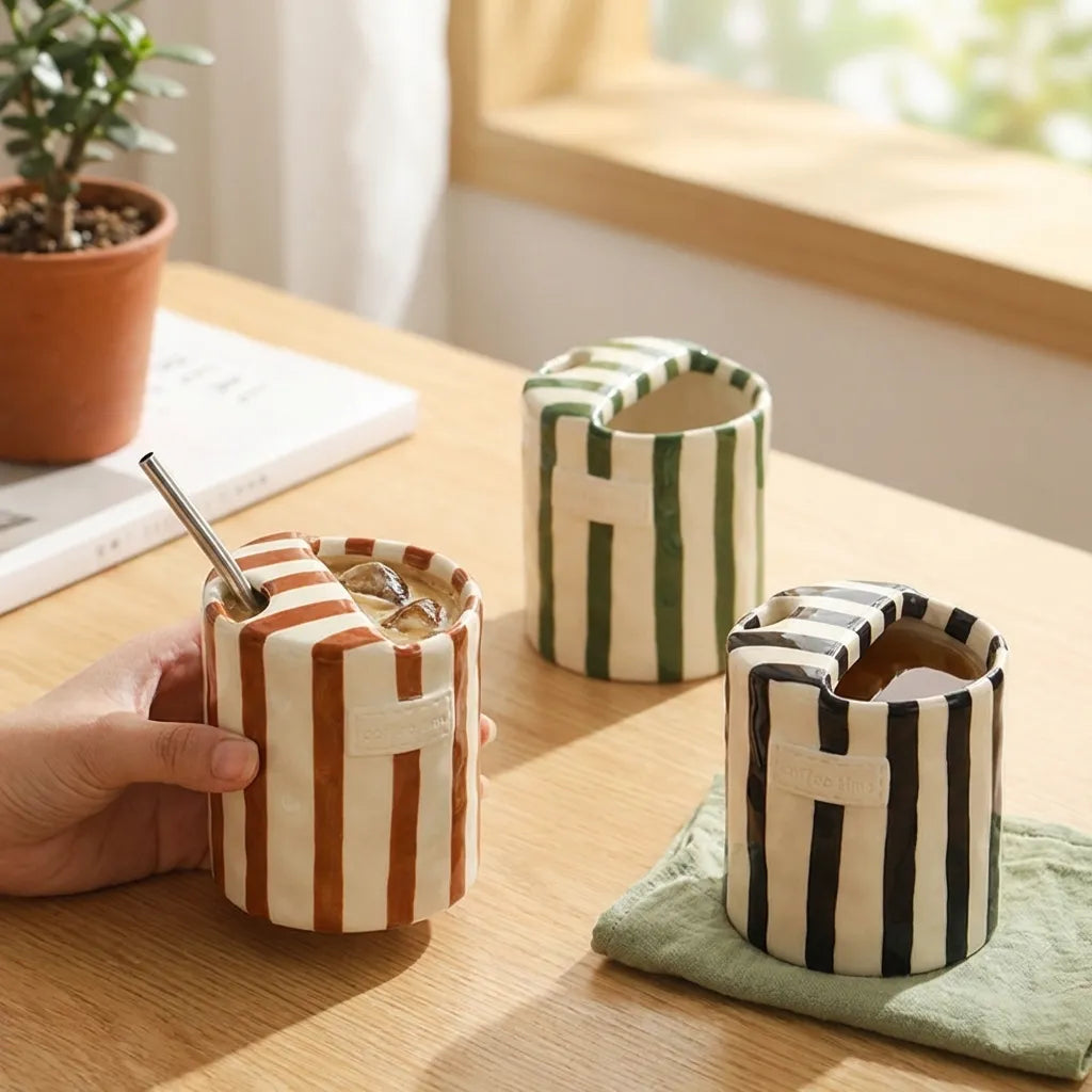 Brown, green and black striped ceramic cups on a desk by a sunny window, one with iced coffee and a straw