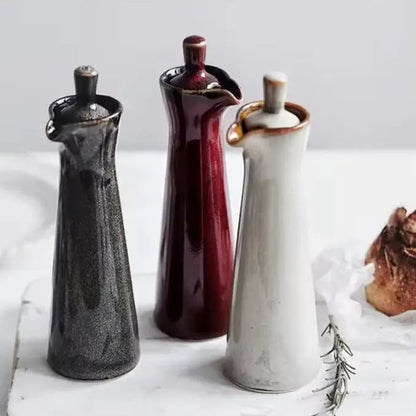Three ceramic oil dispensers in brown, red and beige glazes standing on a white marble board beside a loaf of bread