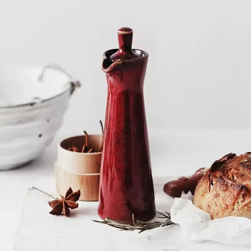 Red ceramic oil dispenser with its stopper in, on a white board beside a loaf of sourdough and star anise