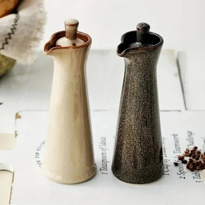Beige and brown ceramic oil dispensers standing side by side on an open cookbook page with peppercorns