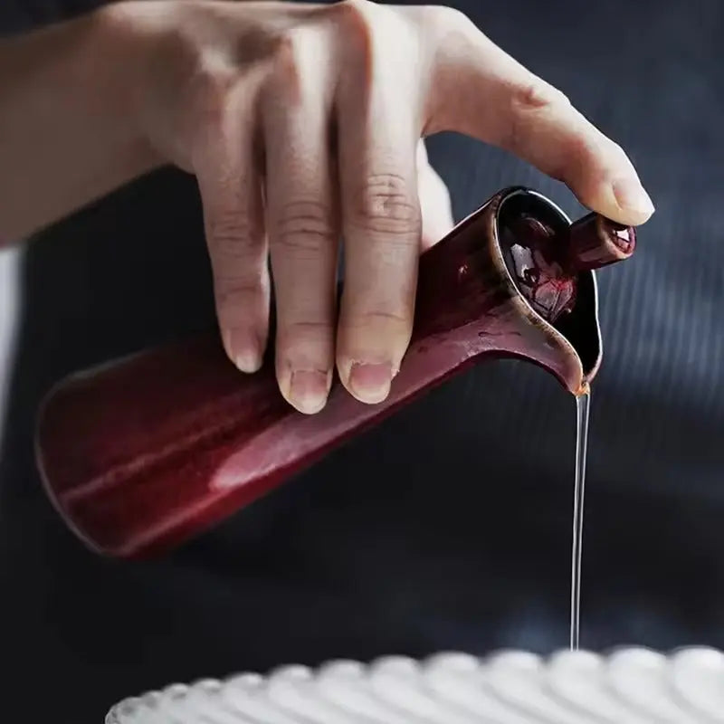 Red ceramic oil dispenser tipped to pour, a thin stream of oil leaving the lip with the stopper still in place