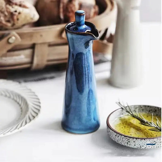 Blue ceramic oil dispenser on a white table with a bread basket behind and a speckled dish of olive oil in front