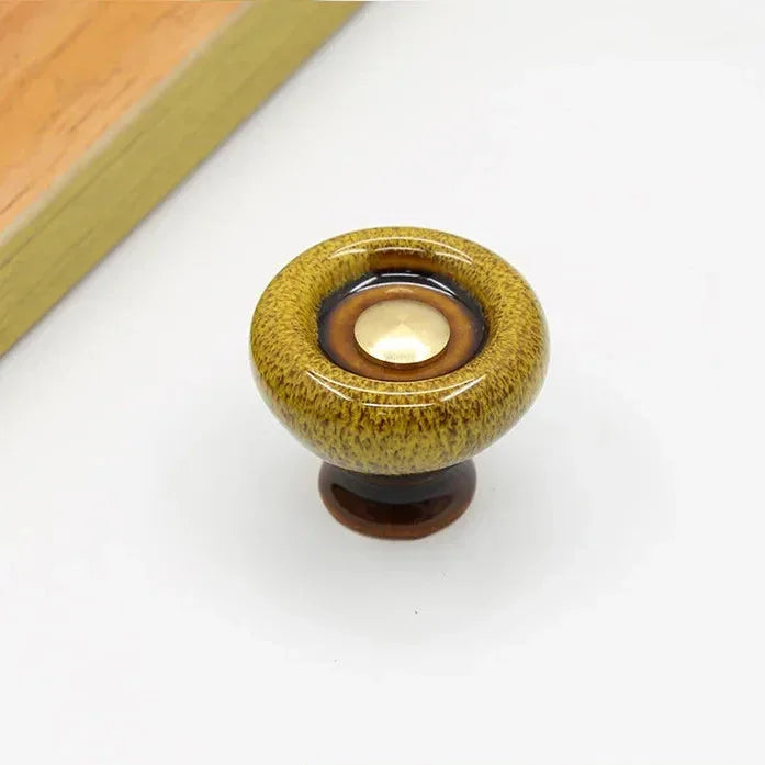 Mustard speckled ceramic cabinet knob with a flared dark brown foot and a dark glaze ring around its gold-tone center cap
