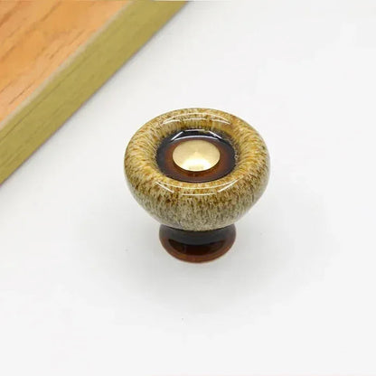 Beige speckled ceramic cabinet knob with a flared dark brown foot and a dark glaze ring around its gold-tone center cap