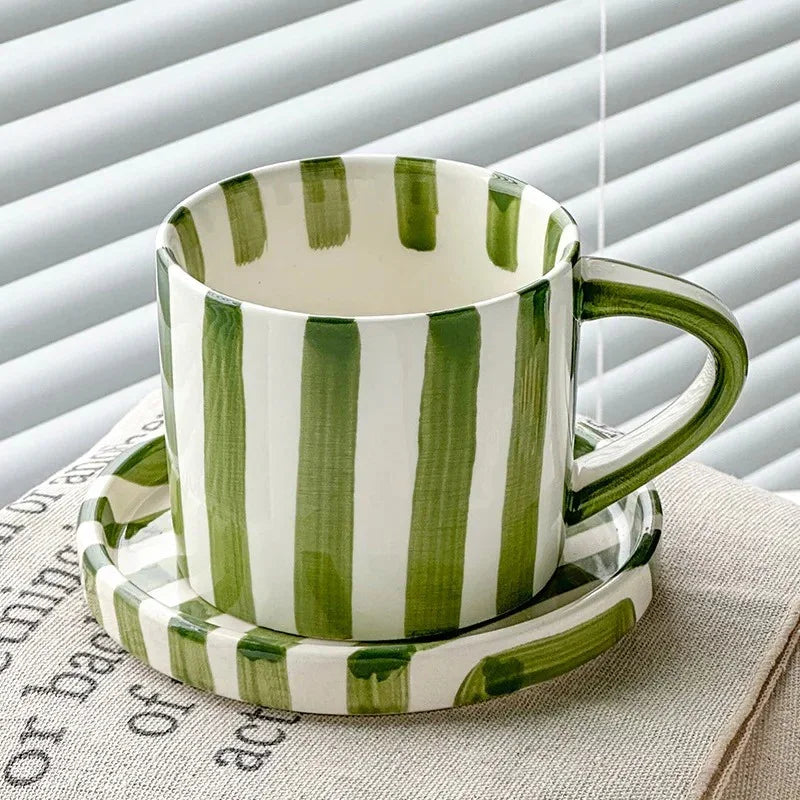 Olive green striped ceramic cup on its matching saucer, by a window with blinds