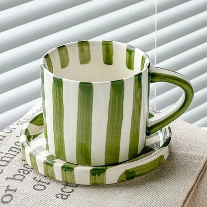 Olive green striped ceramic cup on its matching saucer, by a window with blinds