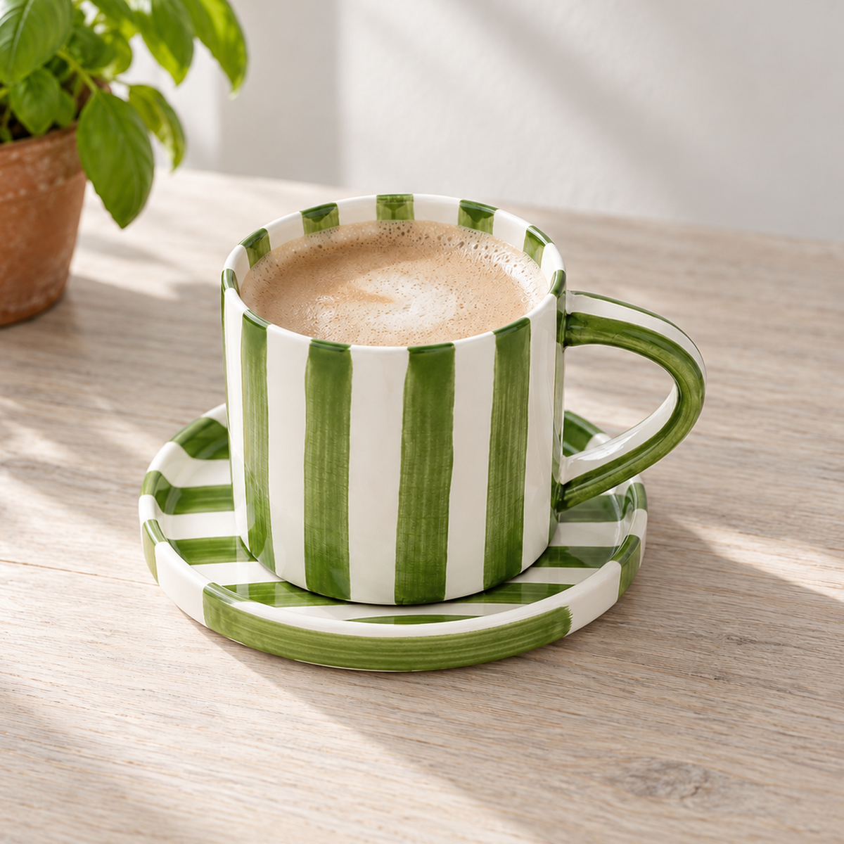 Olive green striped ceramic cup and saucer with milky coffee on a pale wooden kitchen table by a window