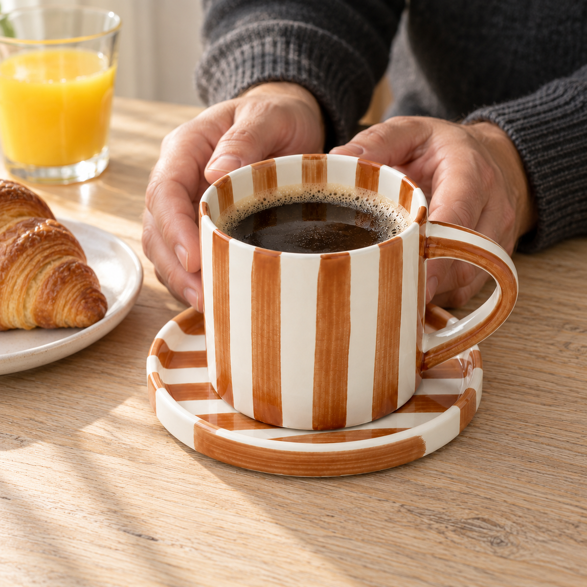 Two hands around a caramel brown striped cup of black coffee on its saucer, with a croissant on a breakfast table