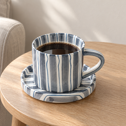 Striped ceramic cup and saucer in slate blue on a small oak side table beside a linen armchair
