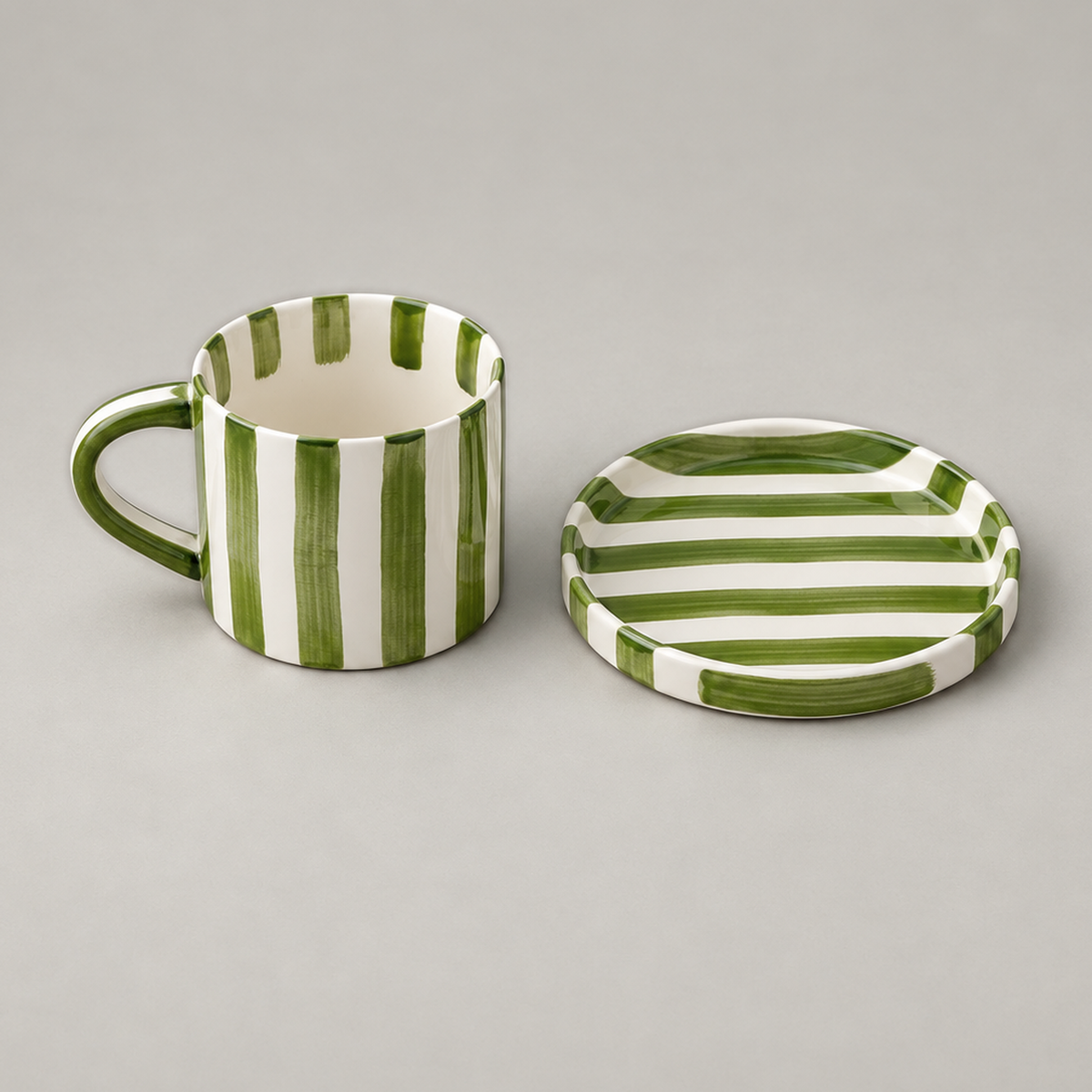 Olive green striped ceramic cup standing beside its empty flat saucer on a plain grey background