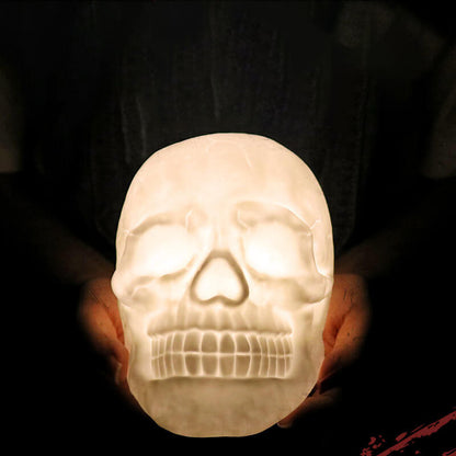 Skull night light glowing warm white, held in two hands in a dark room