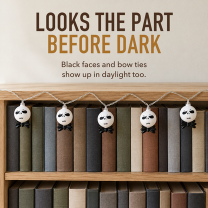Text image: Looks the part before dark. Unlit white string light heads with black faces and bow ties hanging along a light oak bookcase in daylight