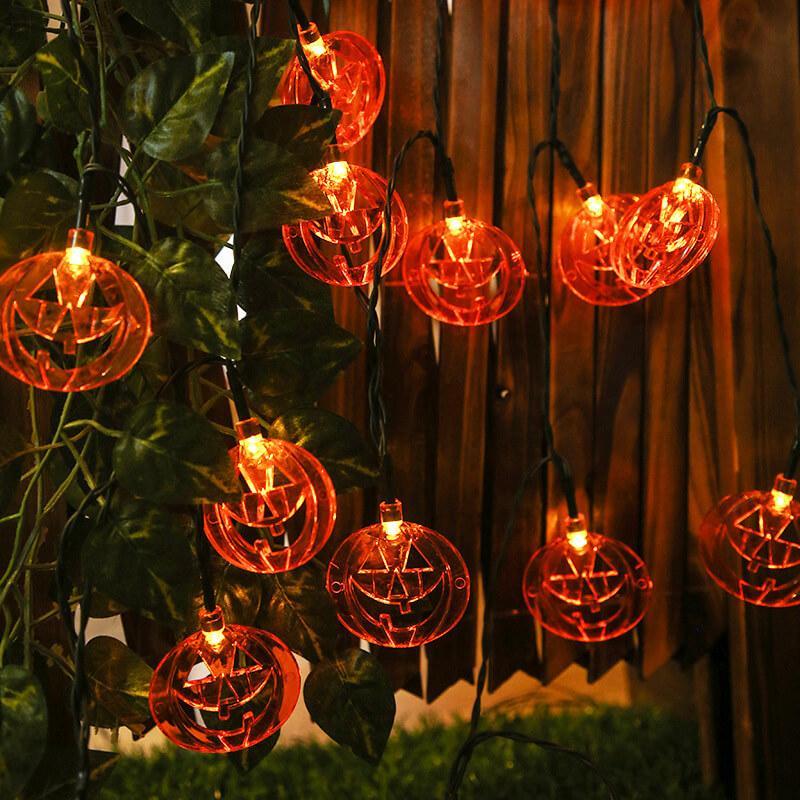 Pumpkin face string lights hung down a wooden fence through ivy, each flat orange pumpkin glowing with its face cut through