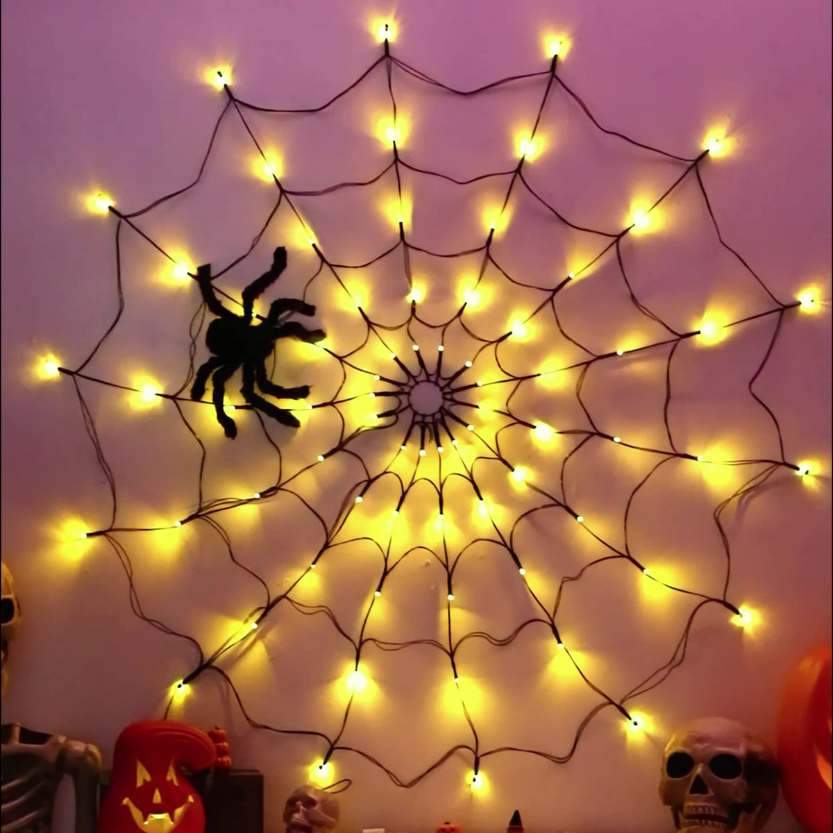 Warm white lit spider web light on a mauve wall with Halloween props below