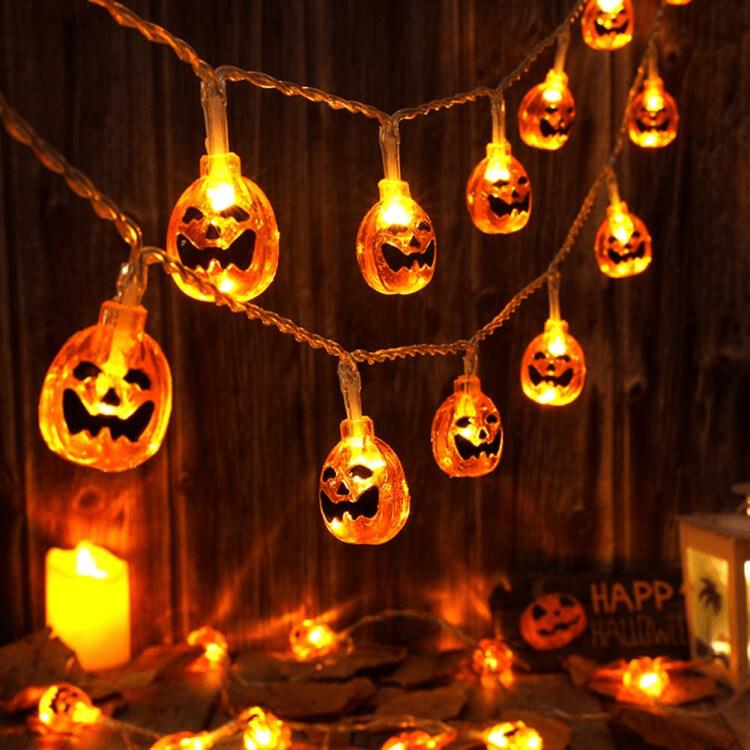 Lit orange jack-o'-lantern string lights on a clear wire swagged across a dark wooden plank wall, with candles below