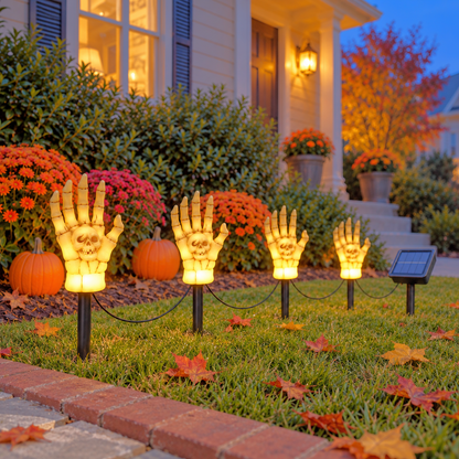 Four skeleton hand stake lights glowing warm in a front lawn at dusk, wired to one solar panel on its own stake