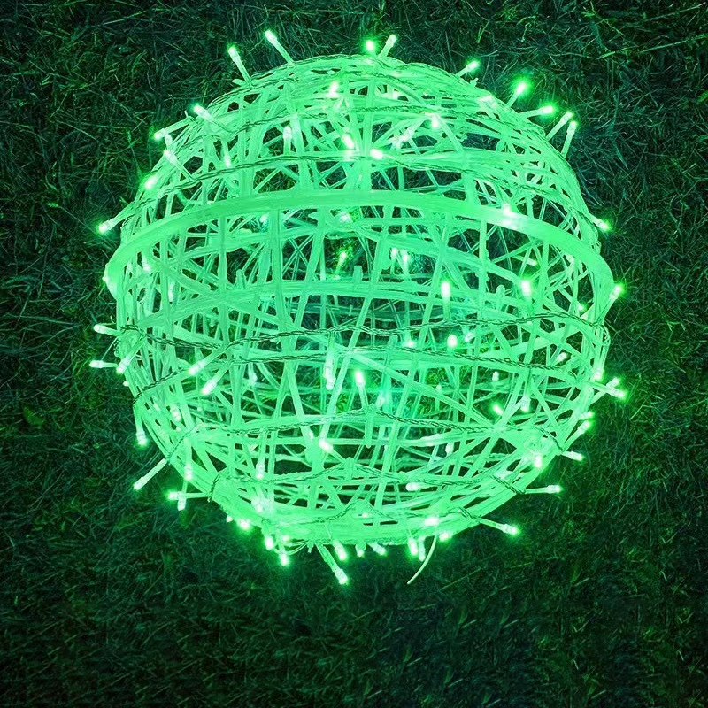 A green LED ball light resting on grass at night, showing its woven strips