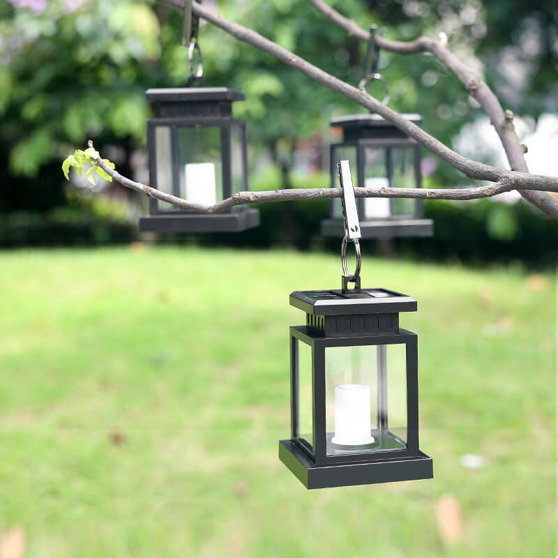 Black solar candle lantern hanging by its metal clip from a branch over a green lawn, with two more lanterns behind