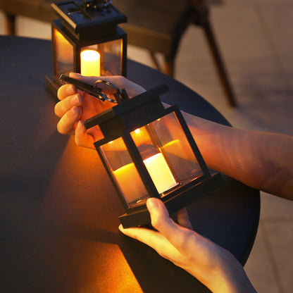 Lit solar candle lantern held in two hands over a table, showing its metal clip and ring