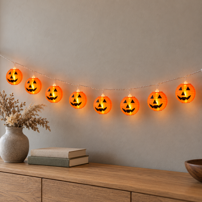 Lit orange lantern string lights with black jack-o'-lantern faces swagged along a pale wall above an oak sideboard in daylight