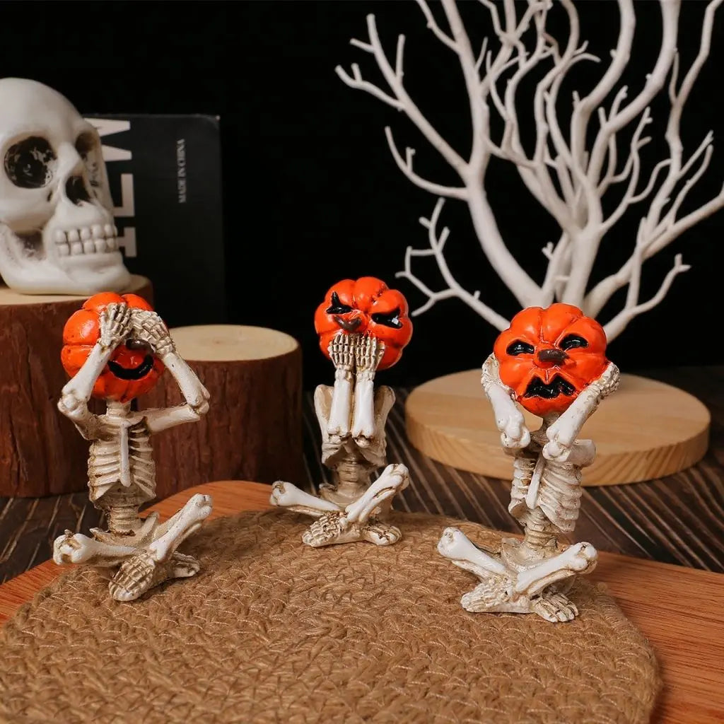 Three pumpkin head skeleton figurines sitting cross-legged in a row on a jute mat, covering their eyes, mouth and ears