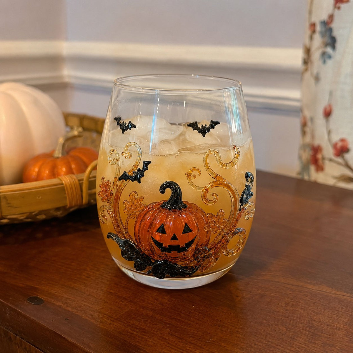 A Halloween drinking glass with a raised pumpkin and bat design, filled with an iced amber drink on a wooden sideboard