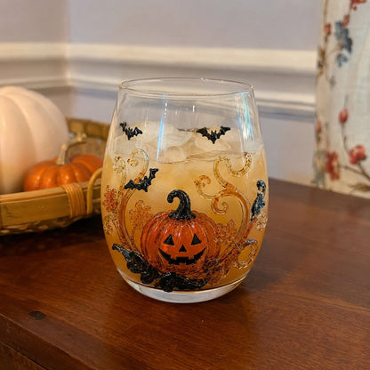 A Halloween drinking glass with a raised pumpkin and bat design, filled with an iced amber drink on a wooden sideboard
