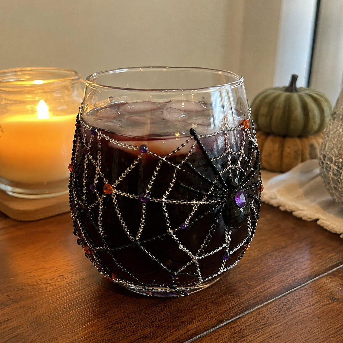 A Halloween drinking glass with a raised beaded spider web design, filled with a dark berry drink beside a candle