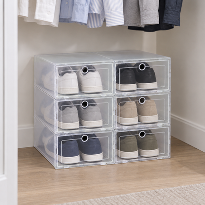 Six clear shoe boxes stacked two across and three high on a closet floor under hanging shirts, a pair of sneakers in each