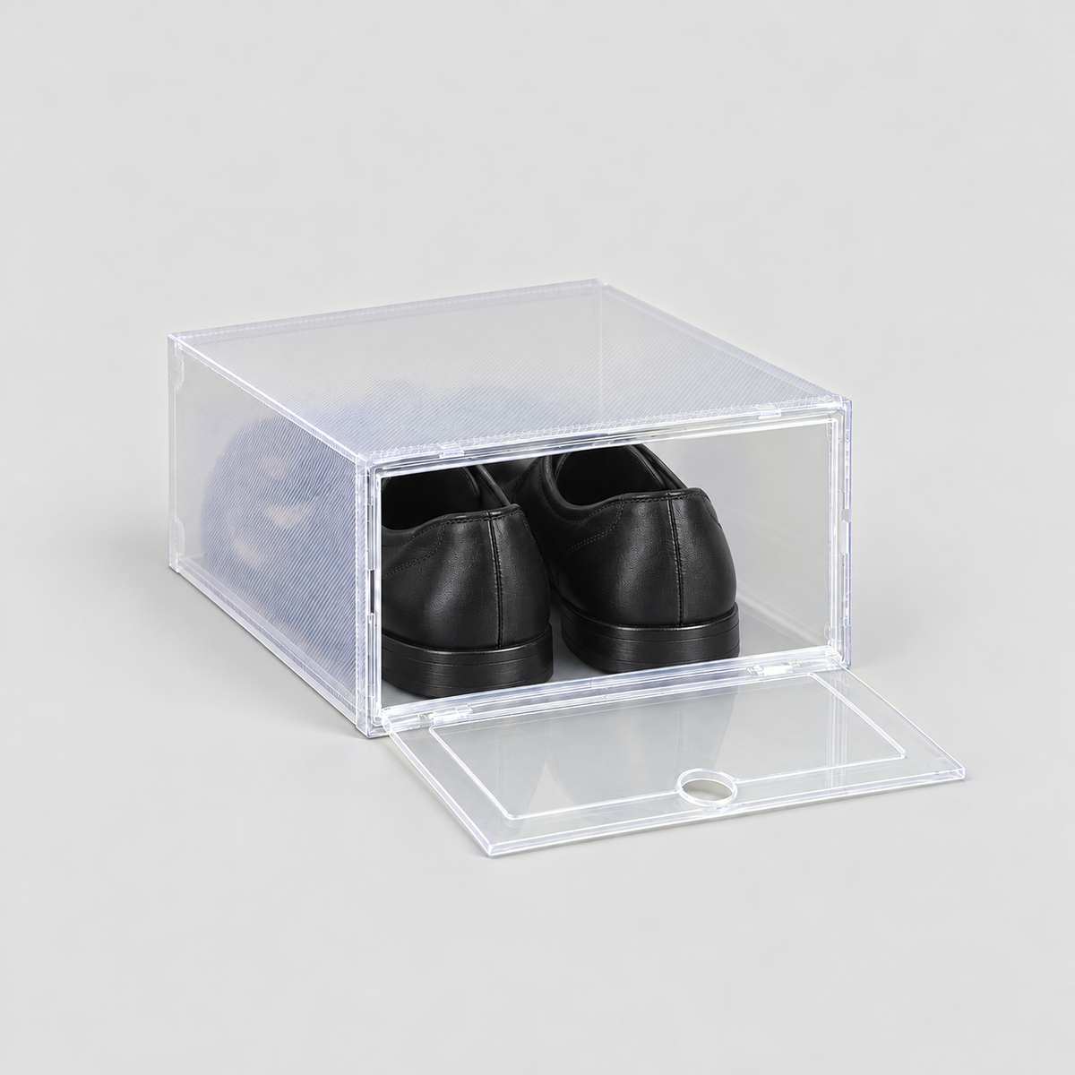 Clear shoe box with its front door dropped open and lying flat, black dress shoes inside, on a plain light grey background
