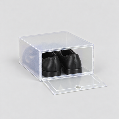 Clear shoe box with its front door dropped open and lying flat, black dress shoes inside, on a plain light grey background