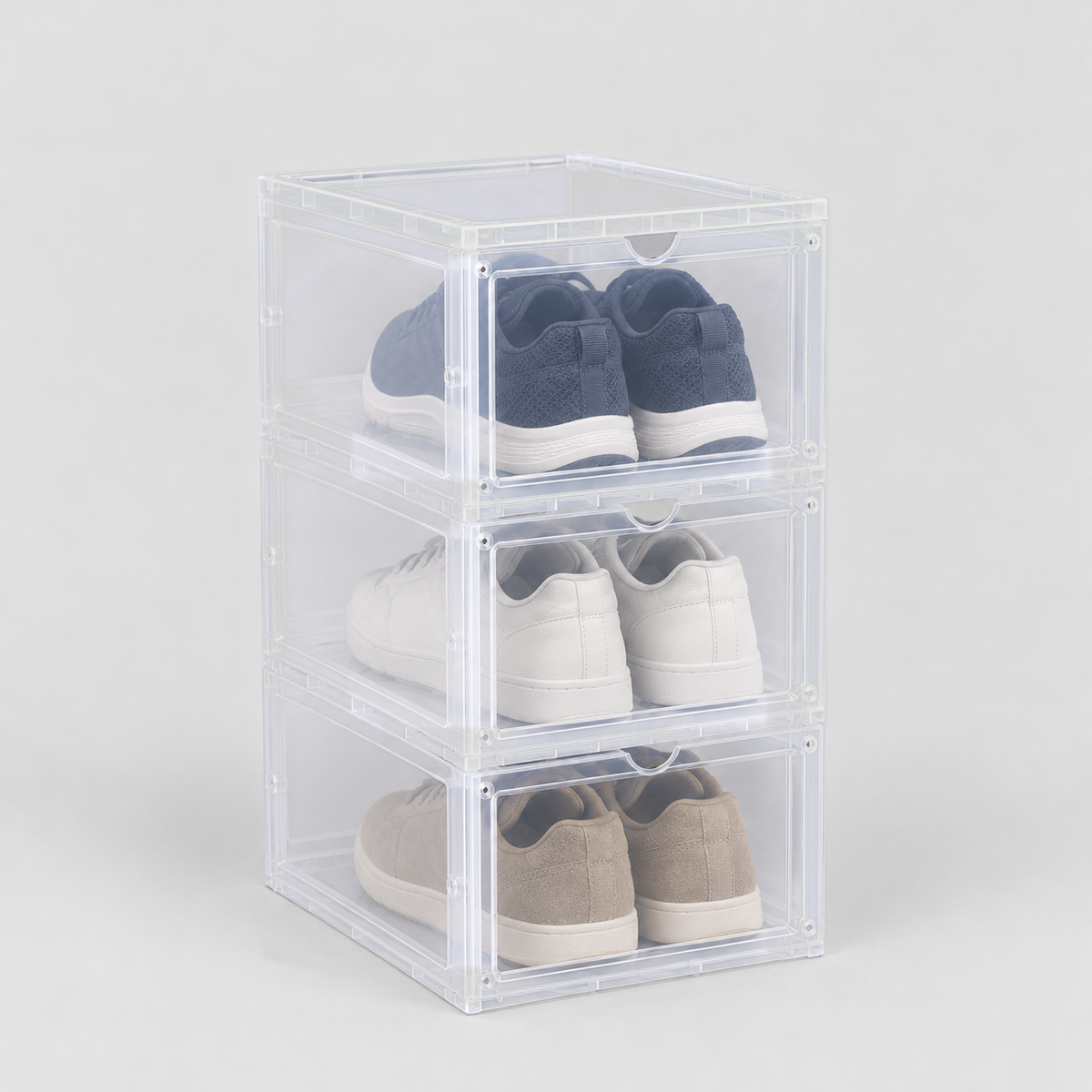 Set of three translucent drop front shoe boxes stacked in one column, a pair of sneakers in each, on a plain light grey background