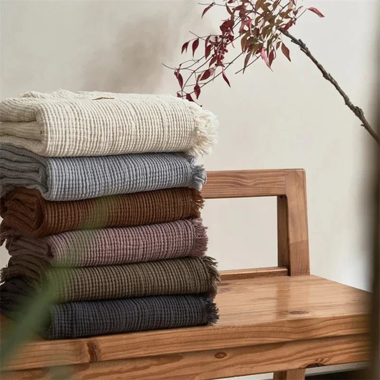 Six folded crinkled cotton throw blankets with fringed edges stacked on a wooden bench, in cream, gray, brown, mauve, olive and charcoal.