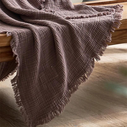 A corner of a mauve crinkled cotton throw blanket with a loose fringe, falling from a wooden bench toward the floor.