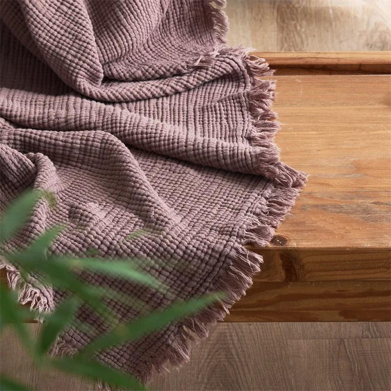 A mauve crinkled cotton throw blanket with a fringed edge draped over a wooden bench.