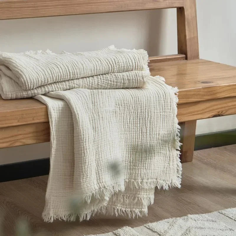 A cream crinkled cotton throw blanket with a fringed edge folded over a wooden bench.